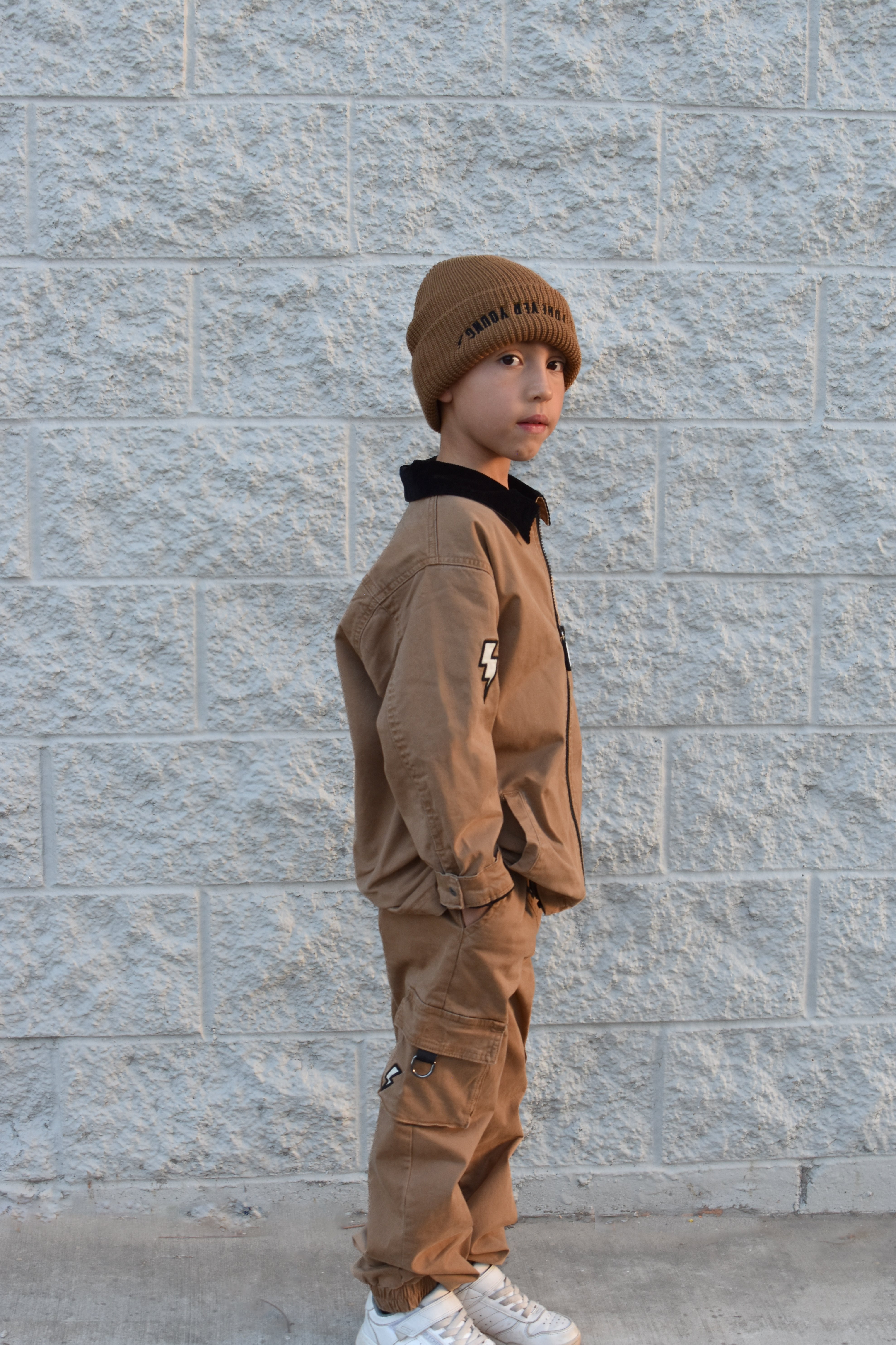 A child wearing a brown beanie with 'Forever Young' text, a matching brown jacket and cargo pants, and white sneakers, standing against a light gray textured brick wall.