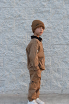 A child wearing a brown beanie with 'Forever Young' text, a matching brown jacket and cargo pants, and white sneakers, standing against a light gray textured brick wall.
