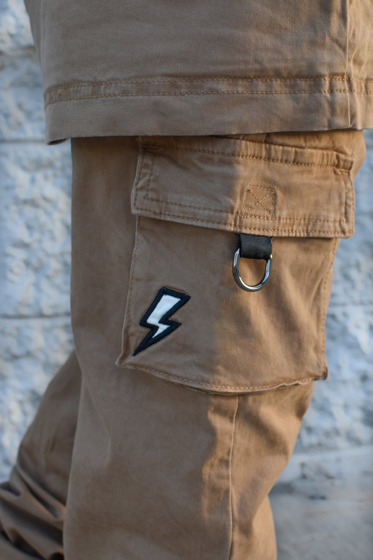 Close-up of tan cargo pants worn by a person, showing a flap pocket with a black-and-white lightning bolt patch and a metal ring detail, against a light textured wall.