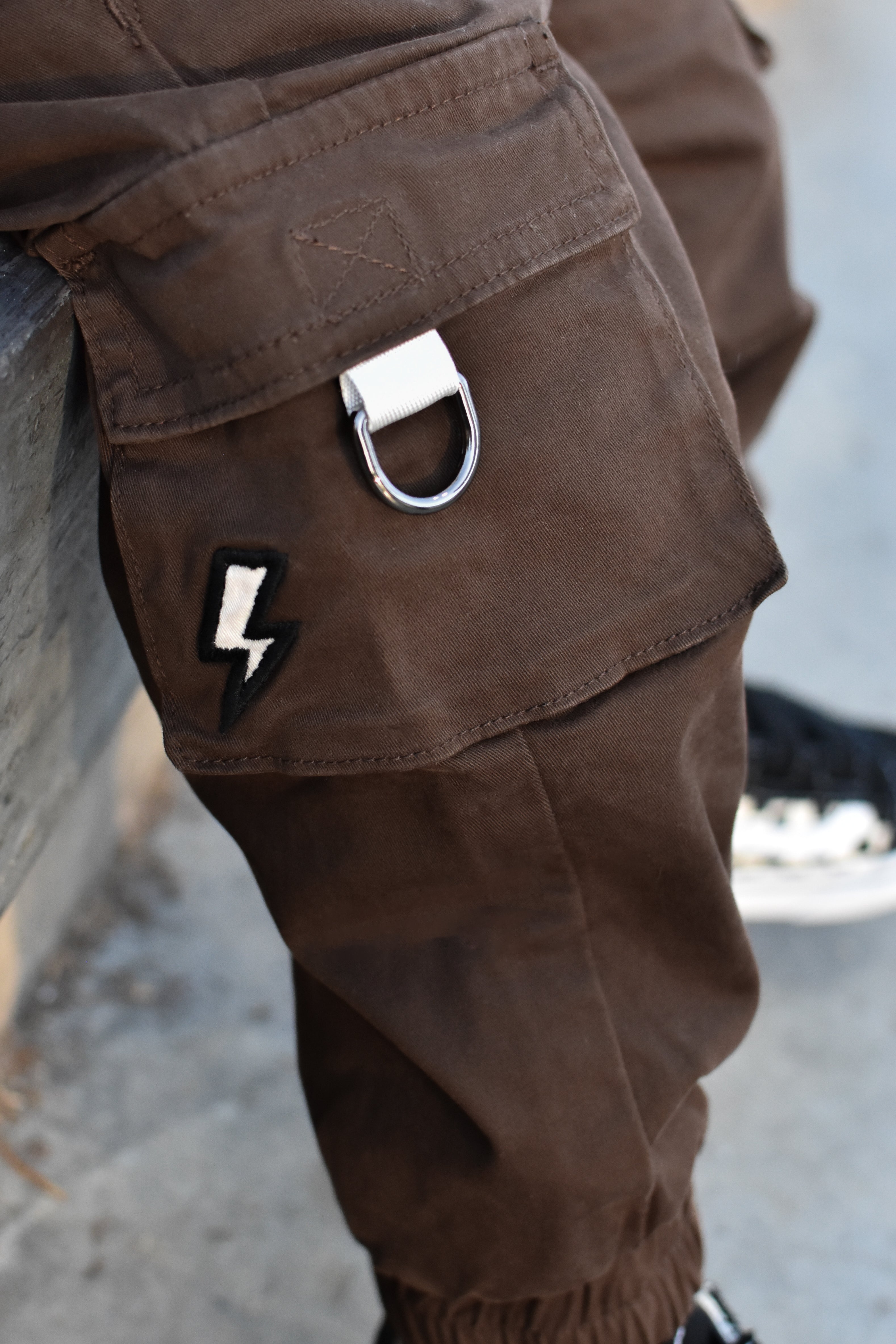 Close-up of brown cargo pants featuring a silver metal D-ring on the cargo pocket and a black-and-white lightning bolt patch.