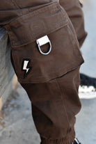Close-up of brown cargo pants featuring a silver metal D-ring on the cargo pocket and a black-and-white lightning bolt patch.