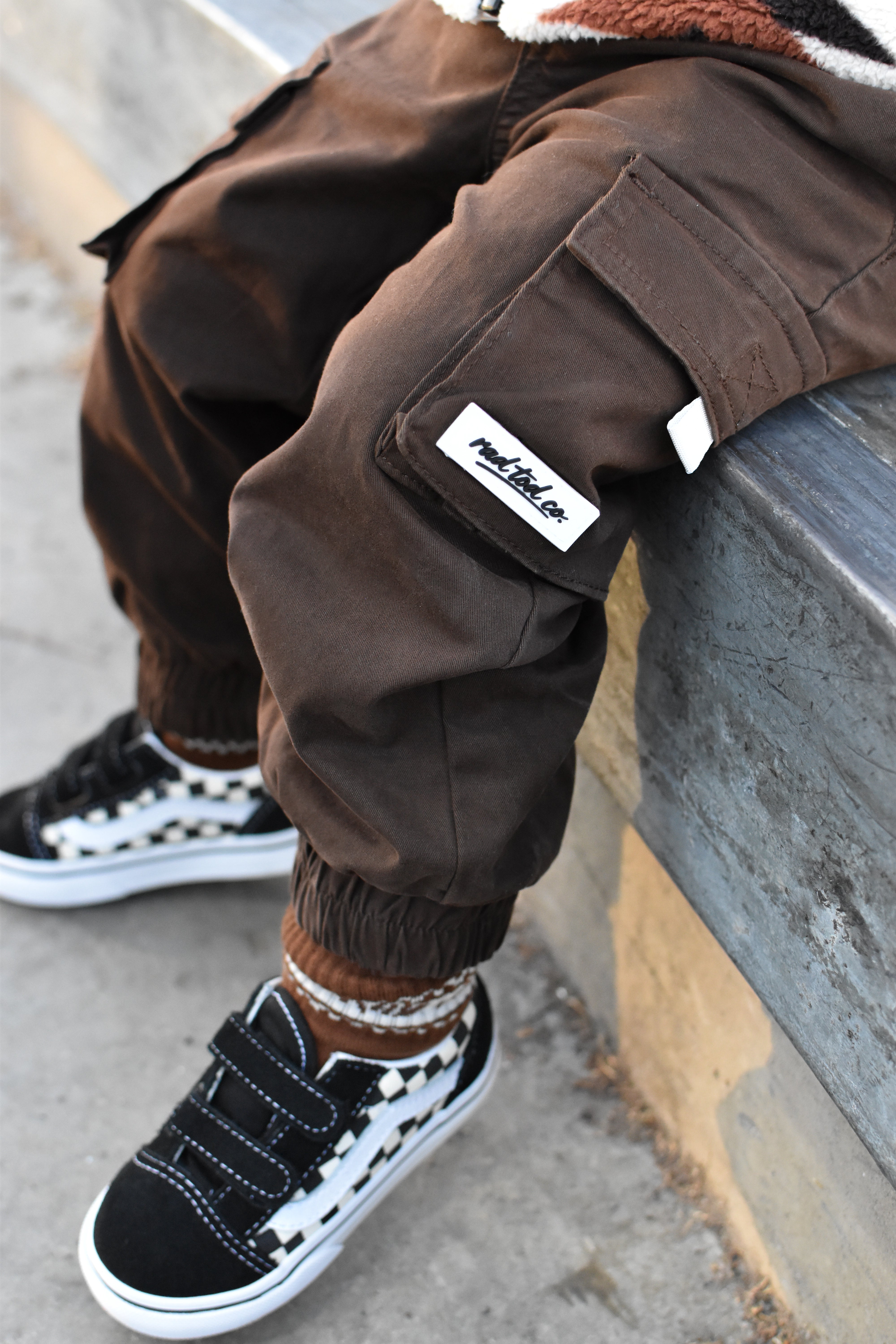 A close-up lifestyle shot of a child wearing brown cargo jogger pants with a visible rad tod co brand patch, paired with black and white sneakers, sitting on a concrete ledge.