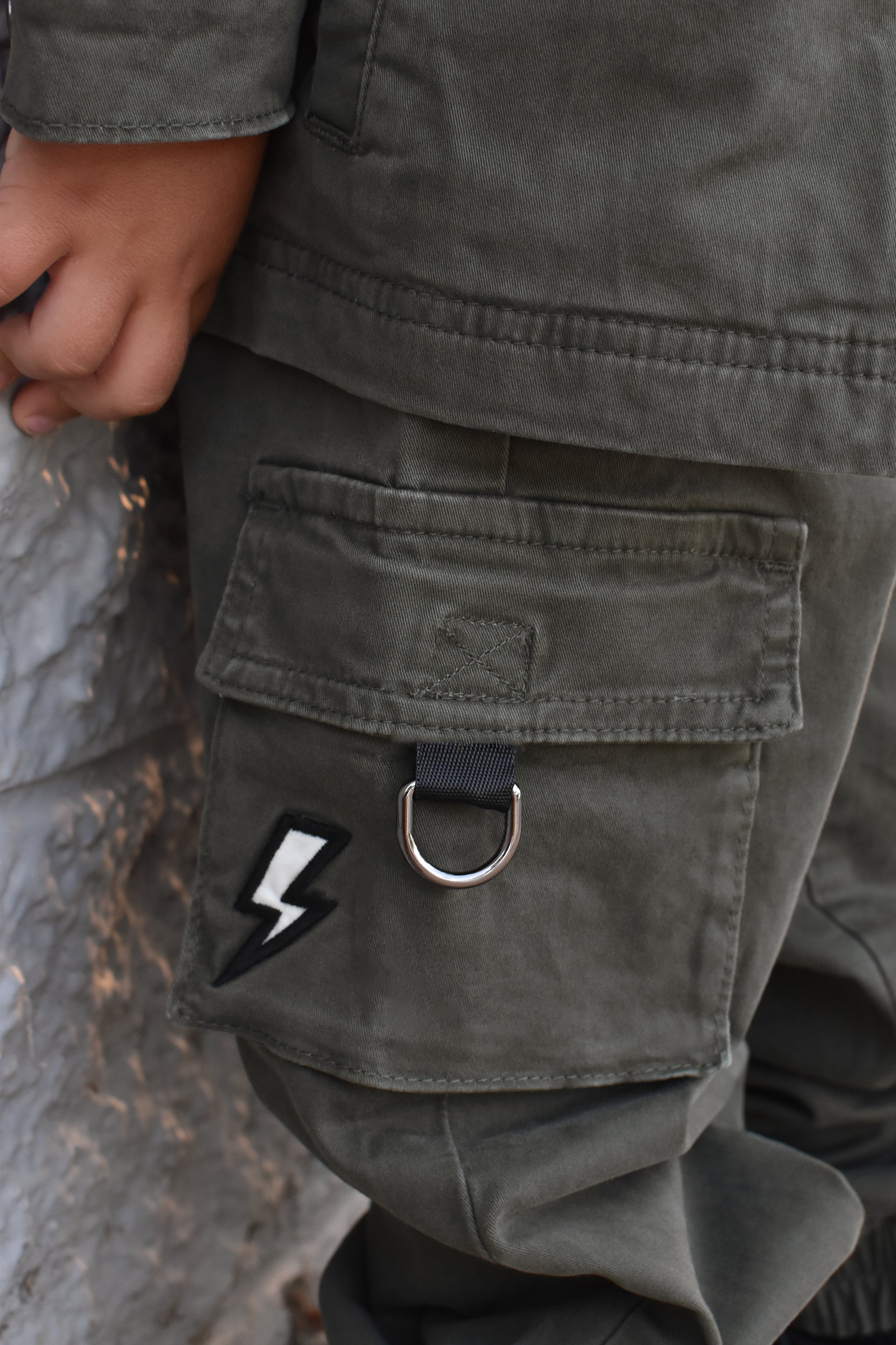 Close-up of a person wearing olive green cargo pants, showing a large front flap pocket with a silver metal D-ring and a black-and-white lightning bolt patch.