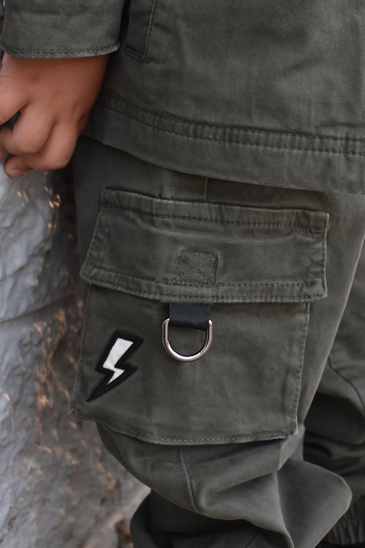 Close-up of a person wearing olive green cargo pants, showing a large front flap pocket with a silver metal D-ring and a black-and-white lightning bolt patch.