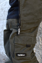 Close-up of olive green cargo pants with a black zipper and a visible 'rad tod co.' brand tab, worn with a black shirt against a light textured wall.