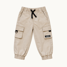 A pair of beige cargo pants for kids, featuring elastic waistband with a black drawstring, side cargo pockets, and elastic cuffs. A small black brand patch is visible on the right pocket flap.