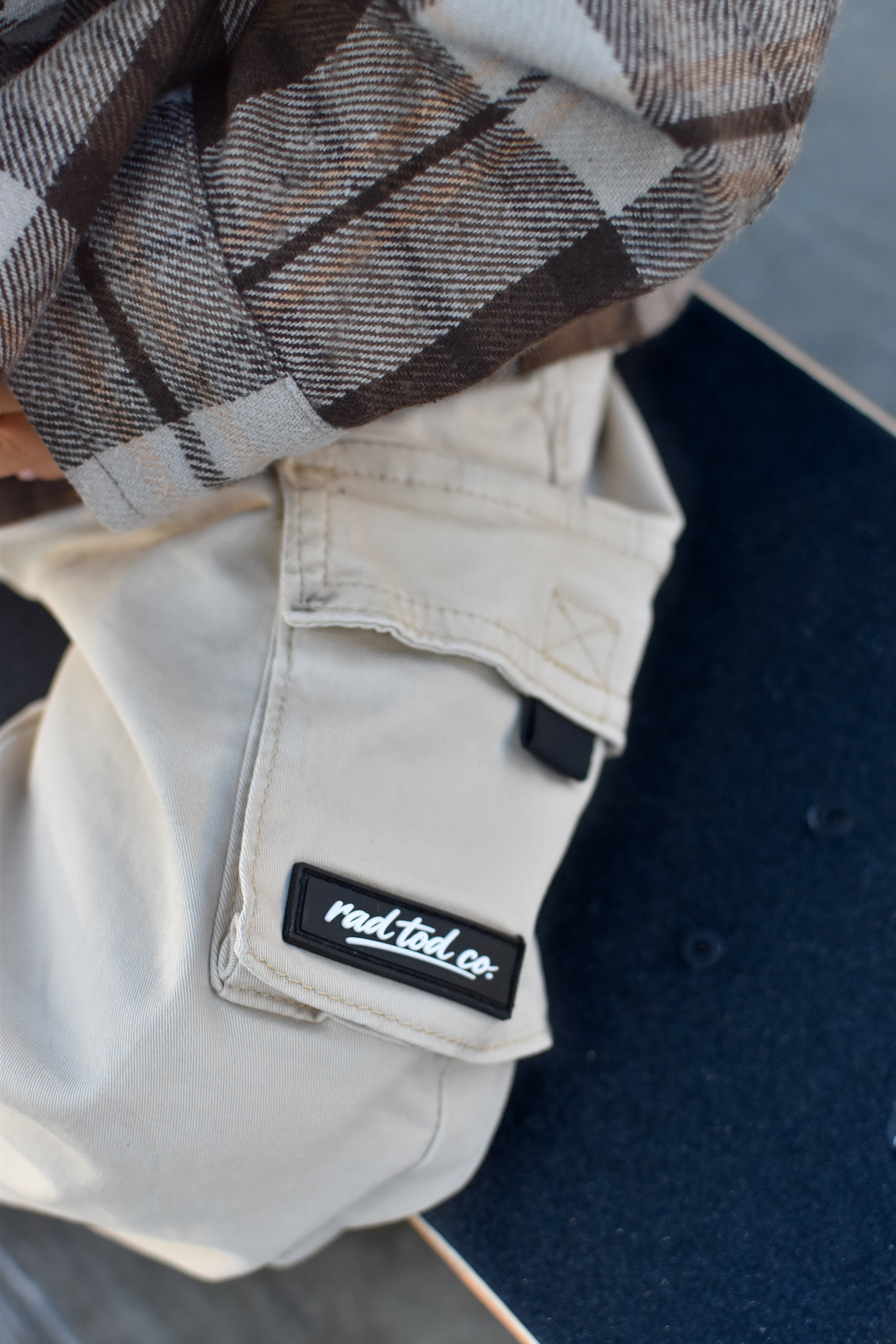 Close-up of a person wearing off-white cargo pants with a black patch that reads 'rad tod co.' and a brown, gray, and white plaid shirt, resting on a dark blue surface.