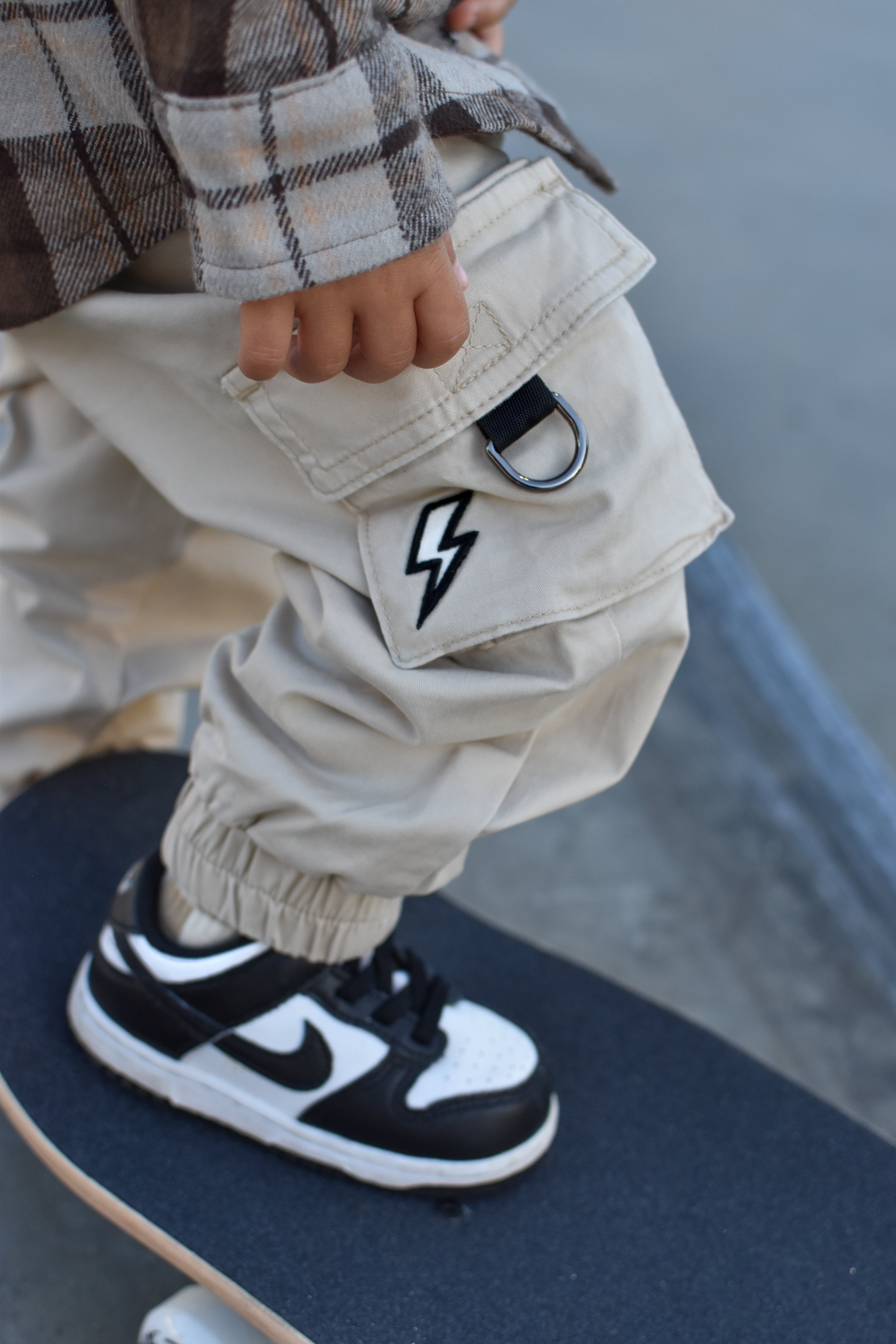 A close-up lifestyle shot of a child wearing beige cargo pants with a black lightning-bolt patch on the pocket, a brown plaid shirt, and a black and white Nike sneaker while standing on a skateboard.