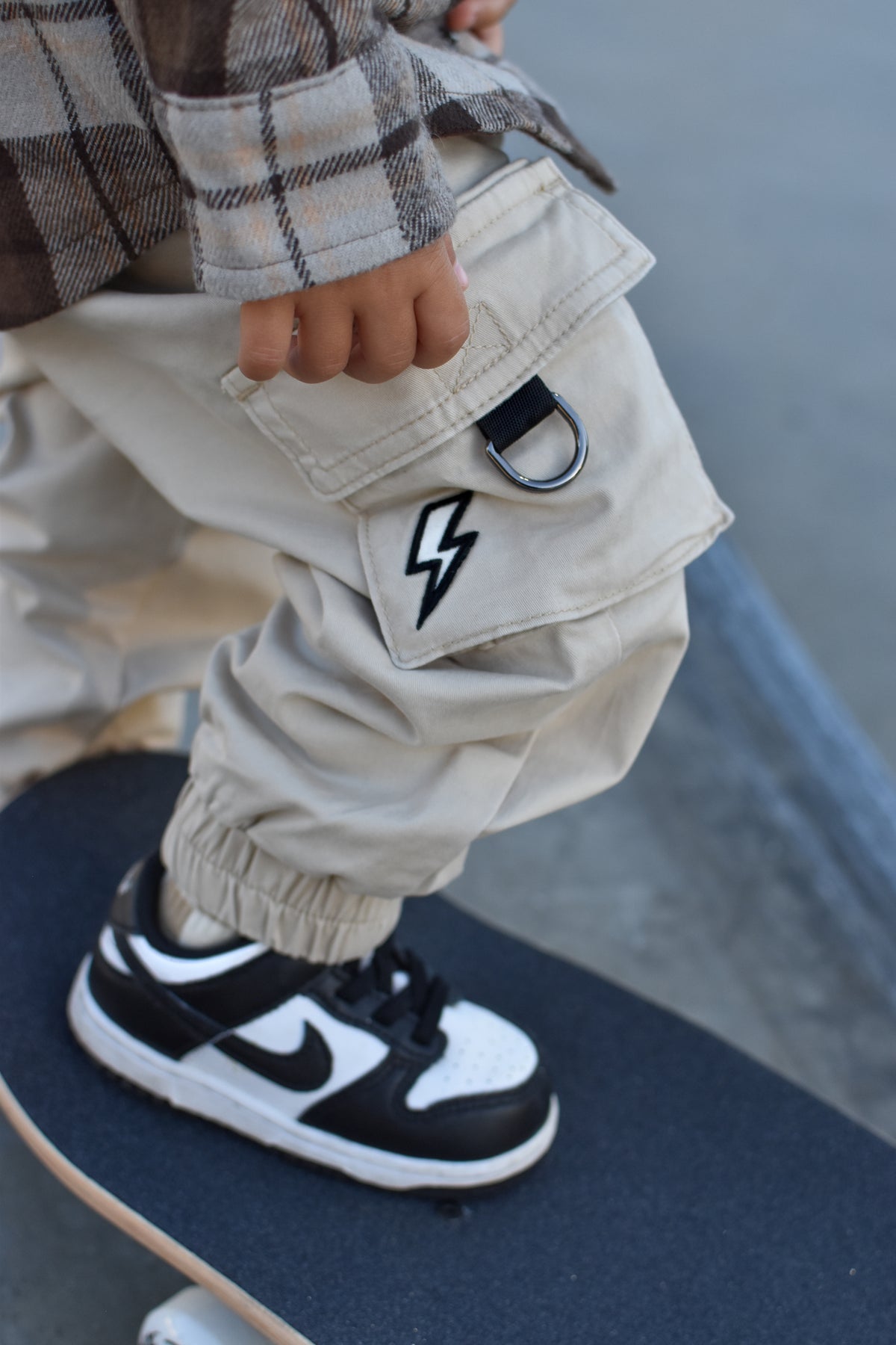 A close-up lifestyle shot of a child wearing beige cargo pants with a black lightning-bolt patch on the pocket, a brown plaid shirt, and a black and white Nike sneaker while standing on a skateboard.