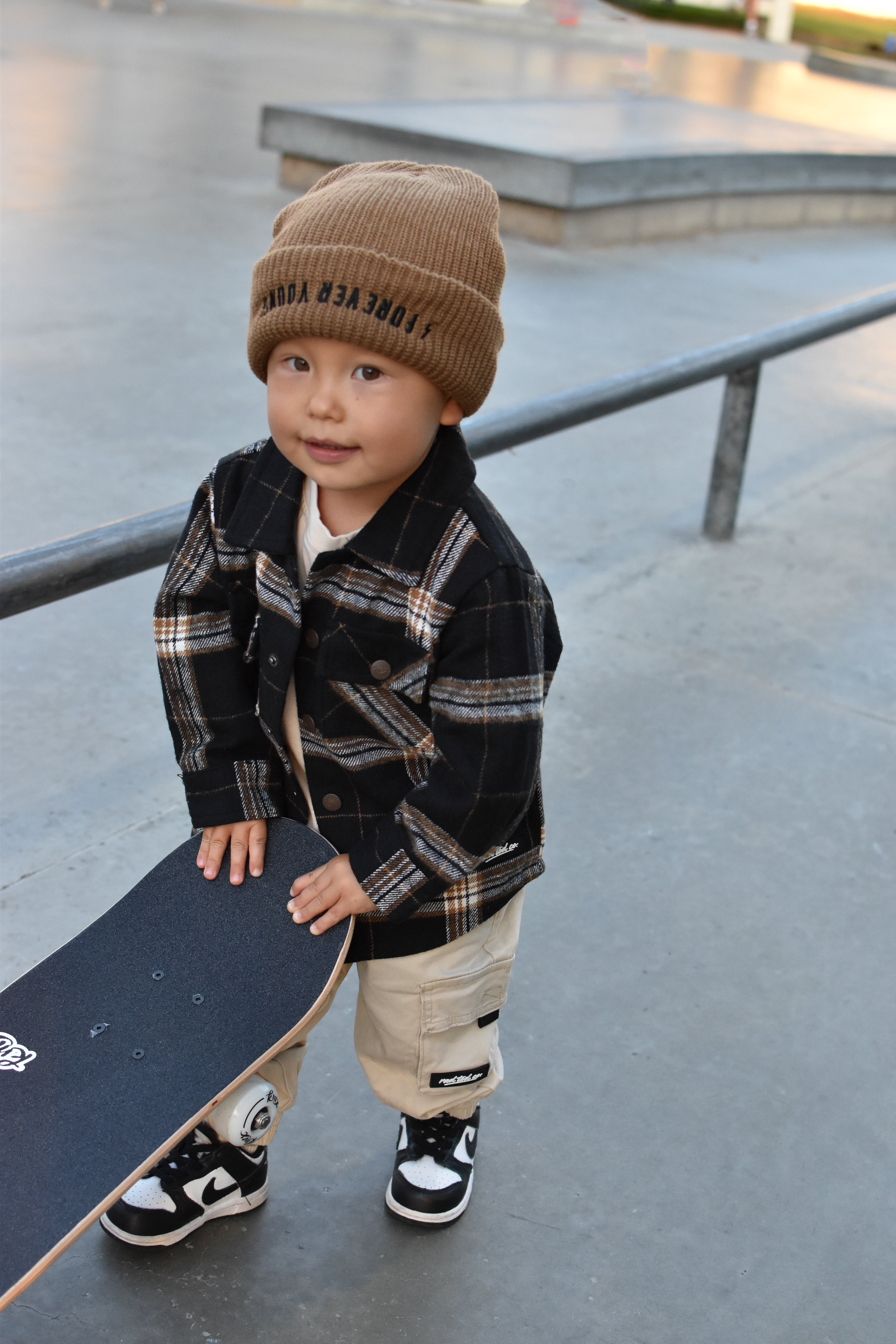 A toddler standing at a skatepark holding a skateboard, wearing a brown knit beanie with black text, a black and brown plaid flannel shirt, beige cargo pants, and black and white sneakers.