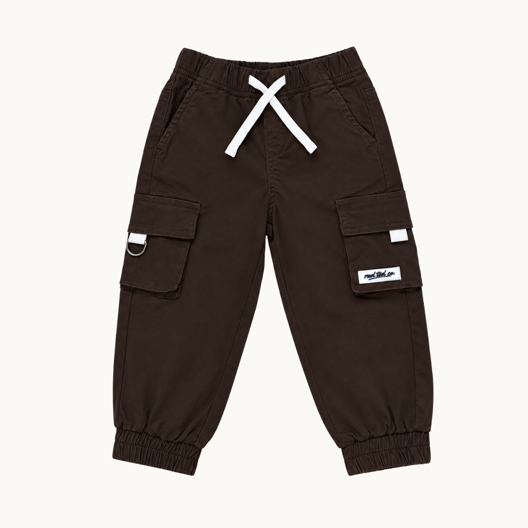 A pair of dark brown cargo pants for kids, featuring a white drawstring, elastic cuffs, and side cargo pockets with small white brand labels, shown against a plain off-white background.