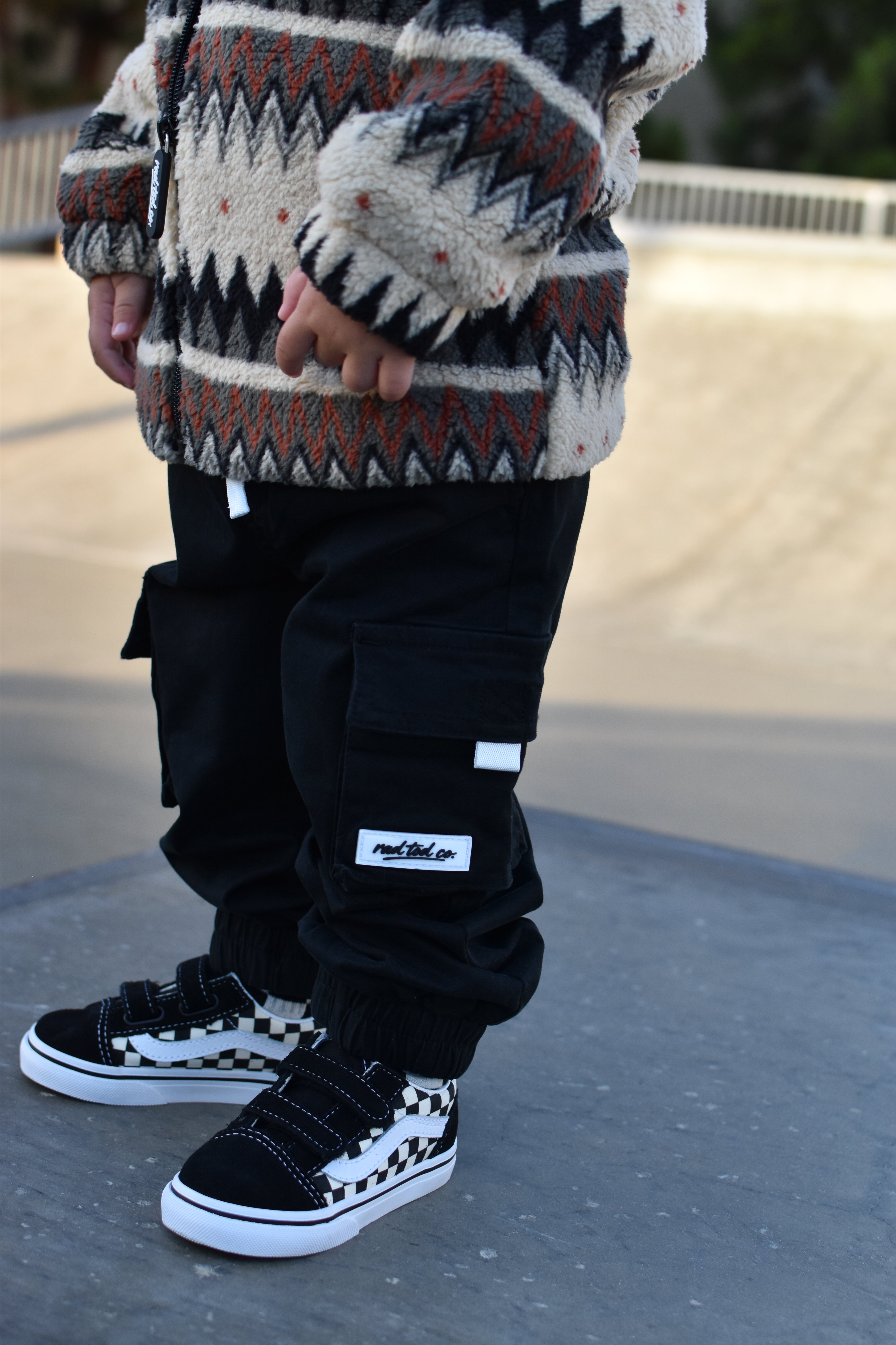 A child wearing black cargo pants with a white 'rad tod co' brand patch, paired with a patterned knit sweater and black-and-white checkered sneakers, standing outdoors on a concrete surface.