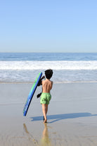 boy walking on a beach with a blue surfboard, wearing lime green shorts by rad toddler