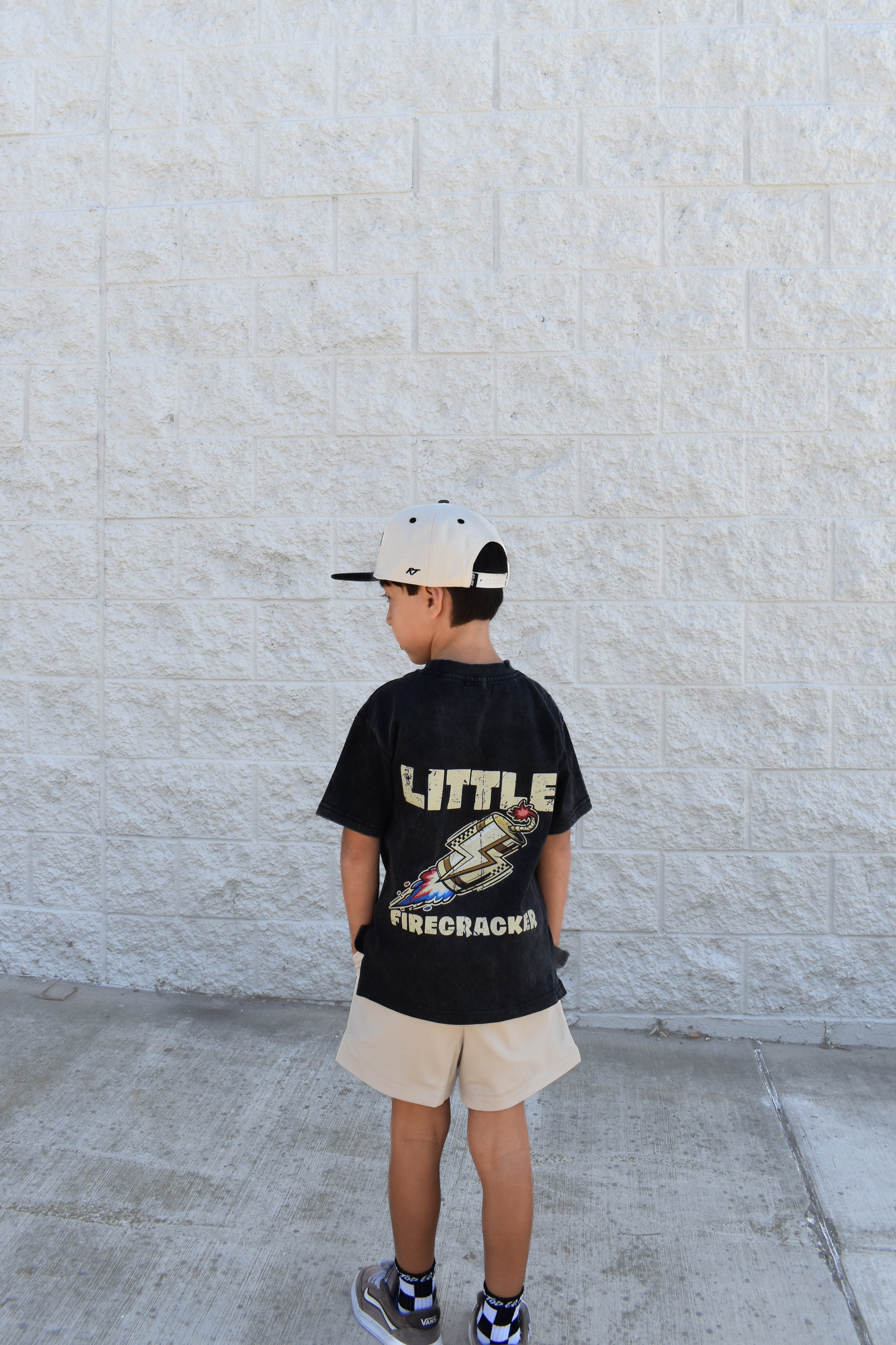 Child wearing a black t-shirt with text and graphics, standing against a light stone wall.
