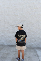 Child wearing a black t-shirt with text and graphics, standing against a light stone wall.