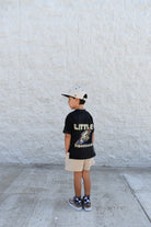 Child wearing a black t-shirt with 'LITTLE FIRECRACKER' text, standing against a light gray wall.