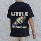 Child wearing a black t-shirt with 'Little Firecracker' design on the back against a stone wall.