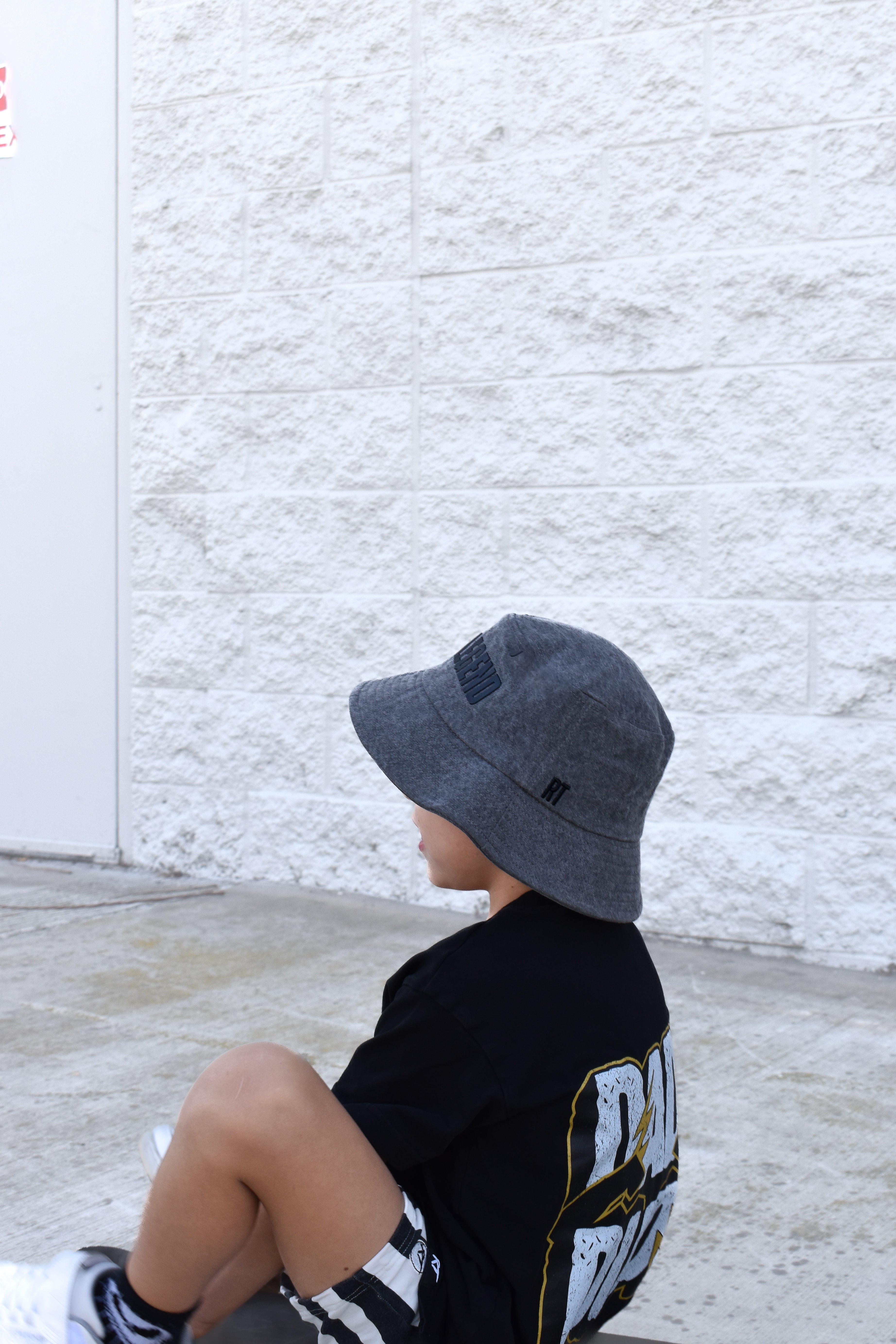 child wearing a gray bucket hat sitting against a textured white wall.