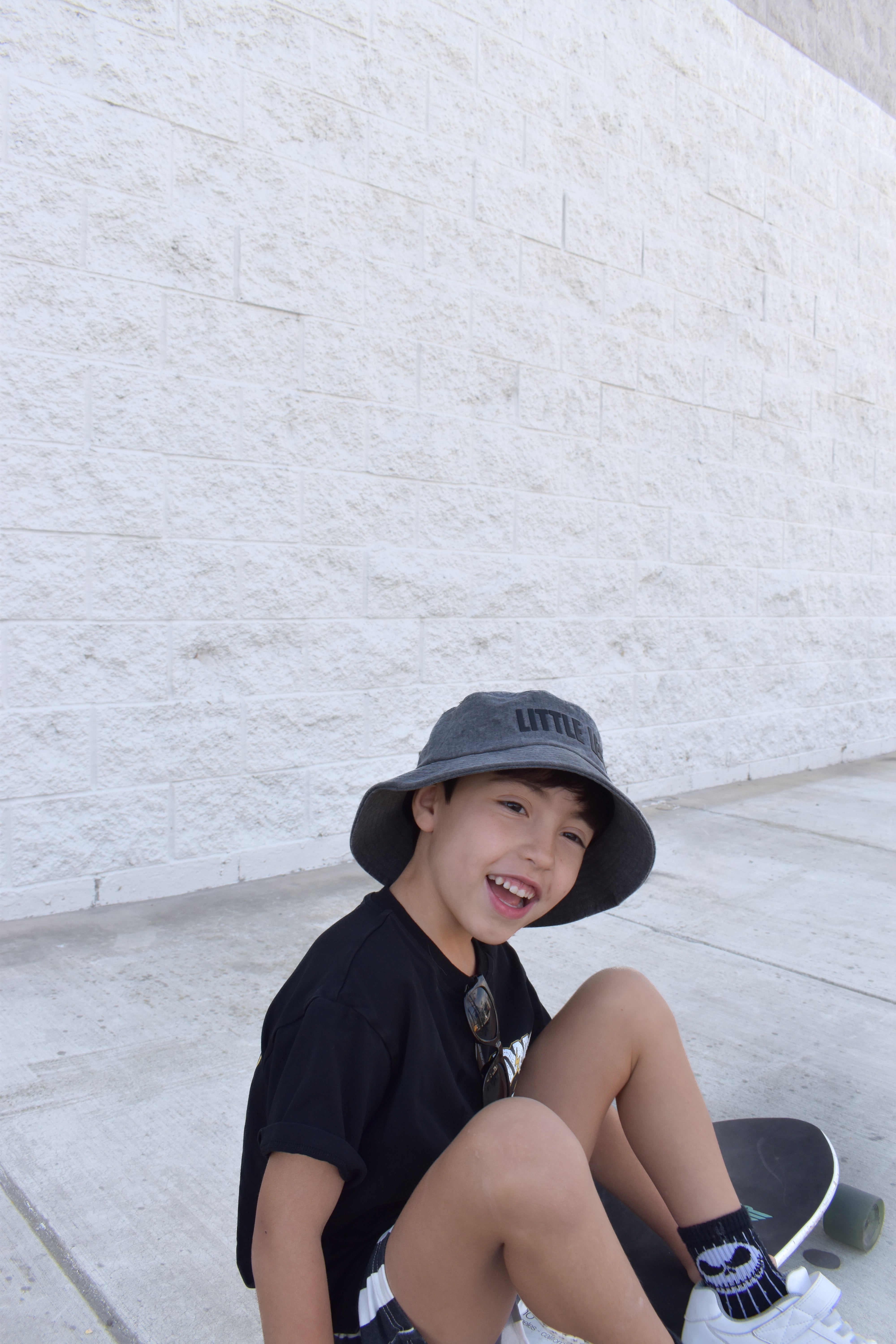 Child sitting on a step wearing a black shirt, gray bucket hat, and white shoes against a light stone wall.