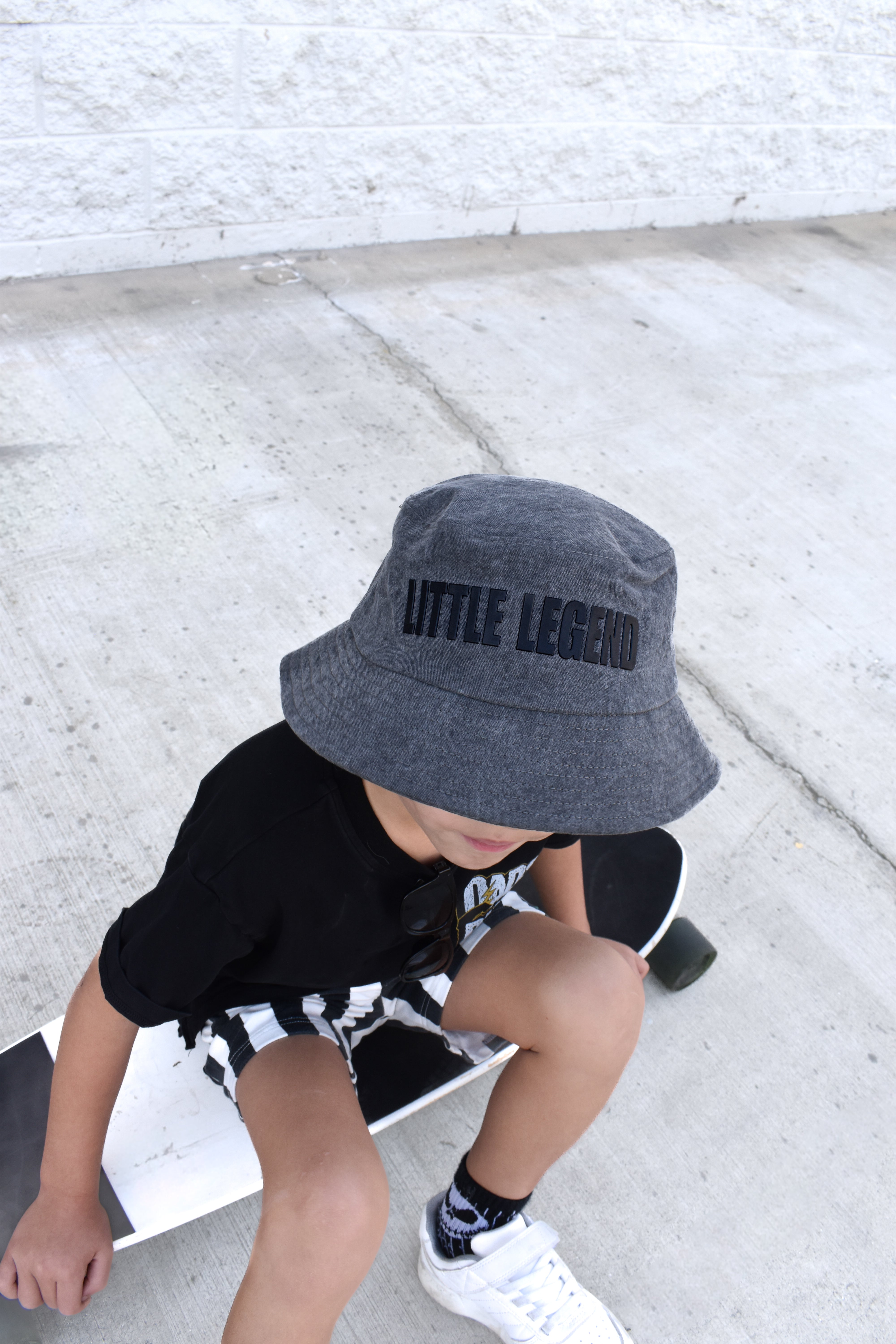 Child wearing a gray bucket hat with 'LITTLE LEGEND' text, sitting on a skateboard.