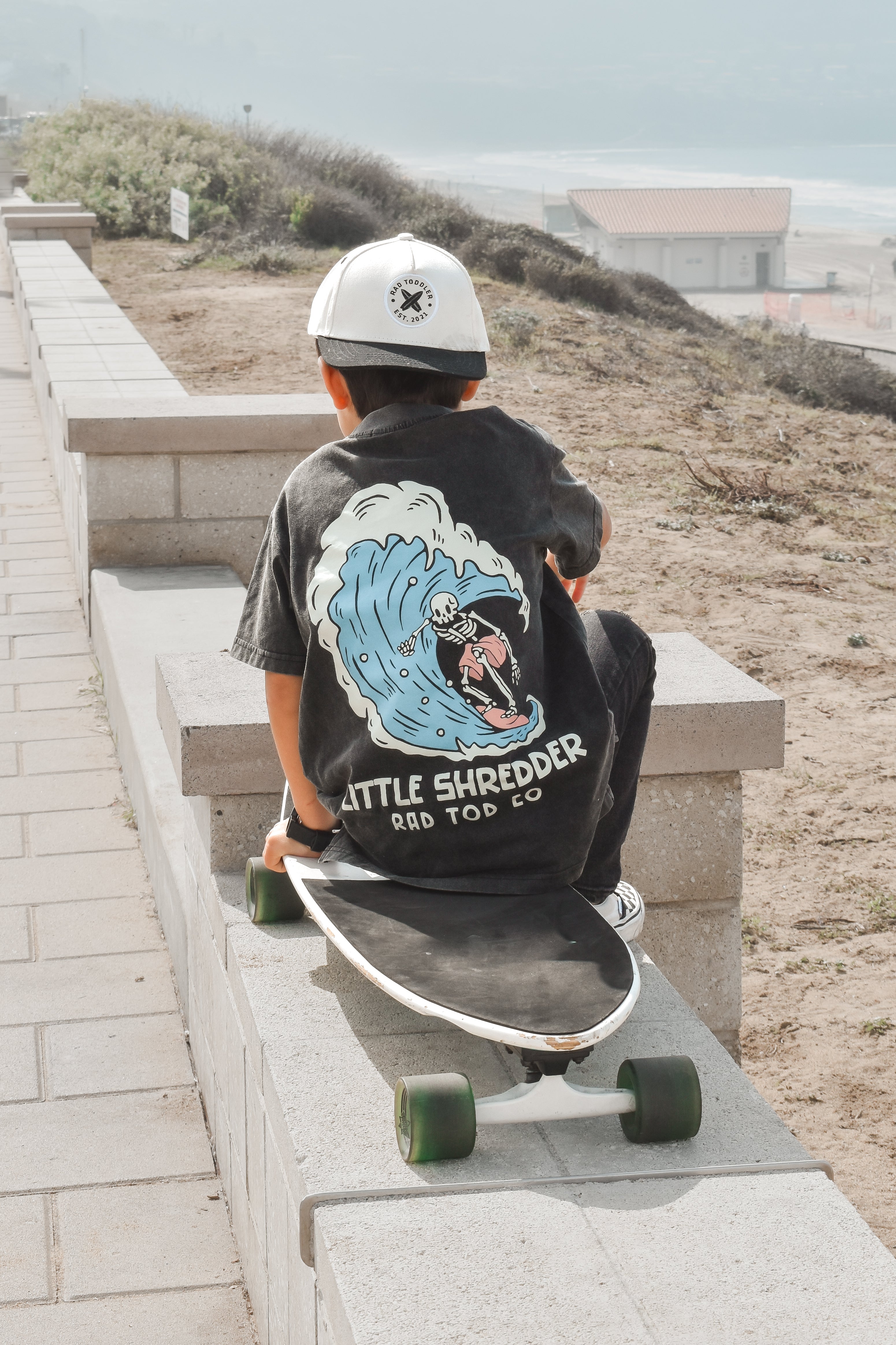 a boy wearing RAD TODS Kids' 'Little Shredder' T-shirt featuring a skeleton surfing a wave design, perfect for young surfers and adventure lovers.