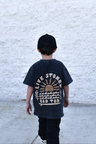 Kid wearing Live Stoked black acid wash t-shirt with golden sun and surfboard design