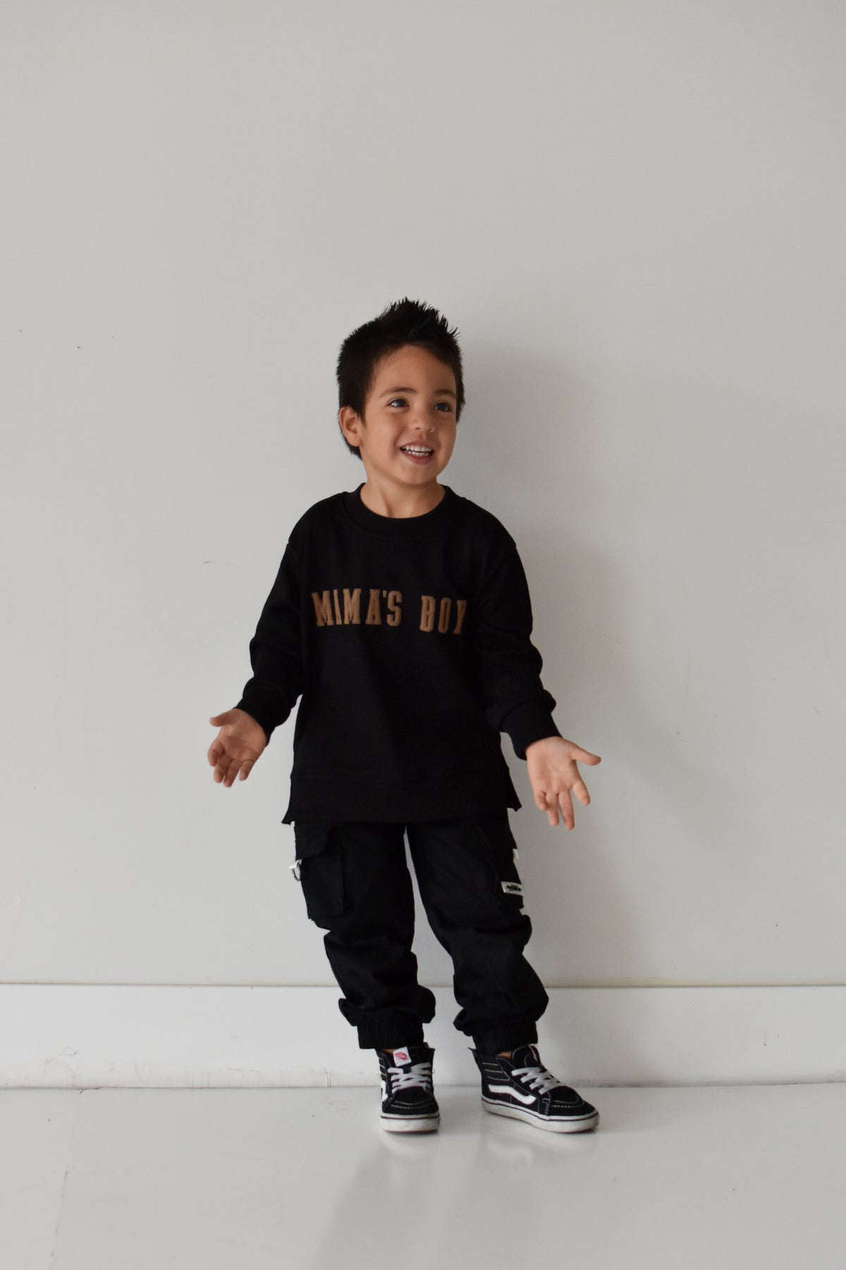 A young boy wearing a black long-sleeve crewneck sweatshirt with 'MAMA'S BOY' printed in gold across the chest, paired with black cargo pants and black sneakers, standing against a plain white wall.