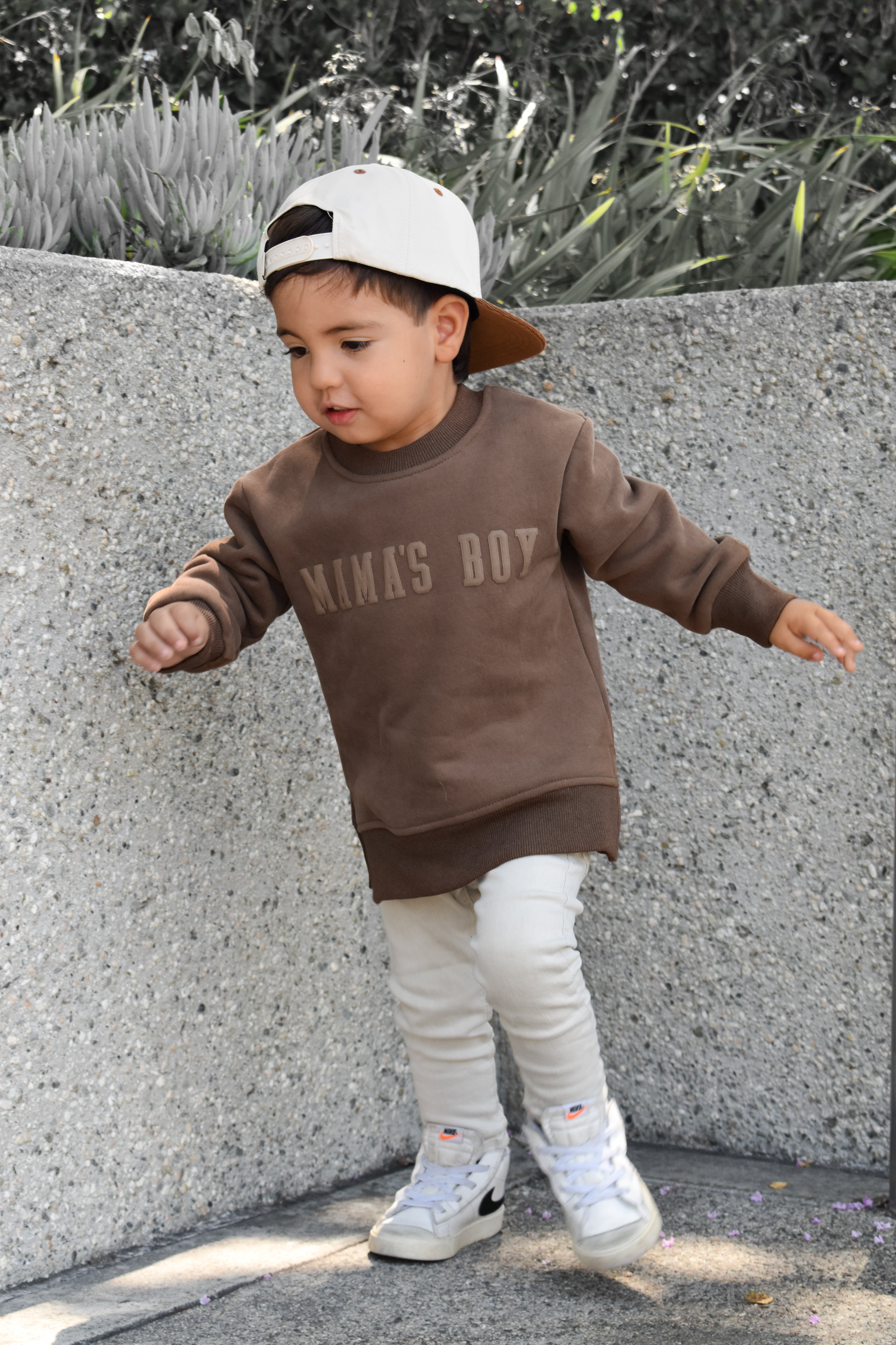 Child wearing a brown 'MamA'S BOY' sweatshirt and white pants standing against a concrete wall.
