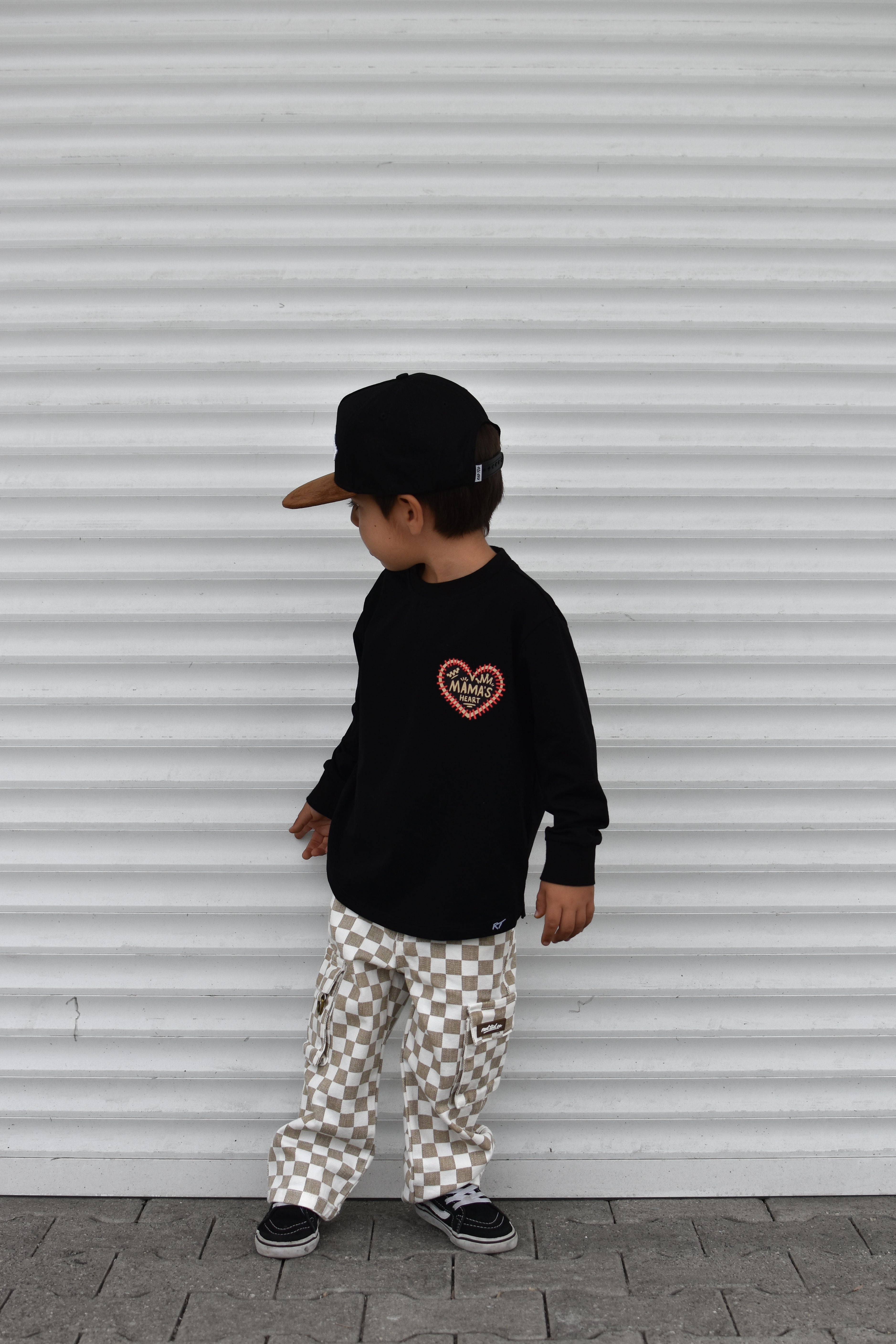 A child wearing a black long-sleeve shirt with a red heart graphic, beige and white checkered cargo pants, a black cap, and black sneakers, standing in front of a white corrugated metal wall.