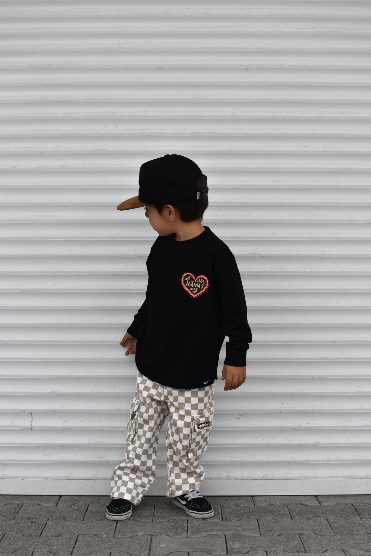 A child wearing a black long-sleeve shirt with a red heart graphic, beige and white checkered cargo pants, a black cap, and black sneakers, standing in front of a white corrugated metal wall.
