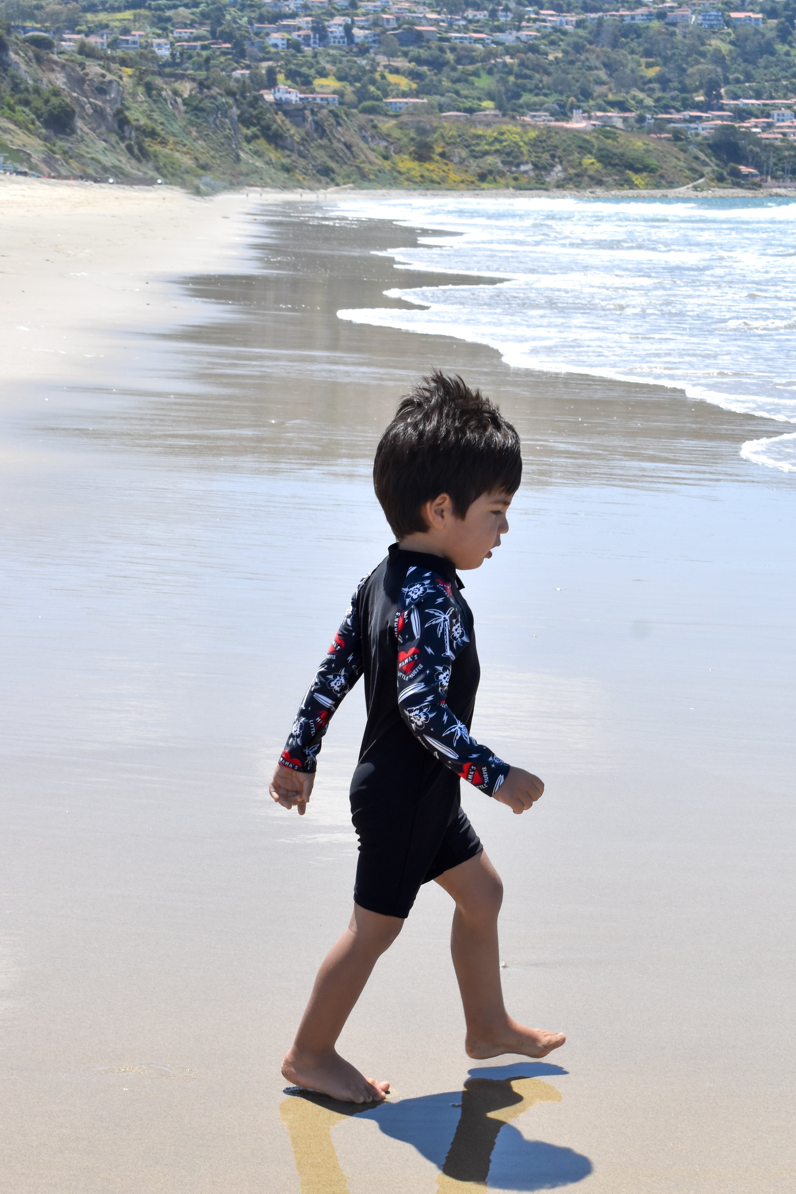Child in a wetsuit walking on a beach with ocean waves and hills in the background