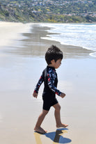 Child in a wetsuit walking on a beach with ocean waves and hills in the background