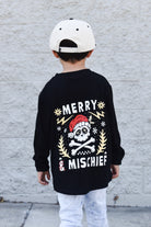 Child wearing a black long-sleeve shirt with a festive design and white cap, standing against a light gray wall.