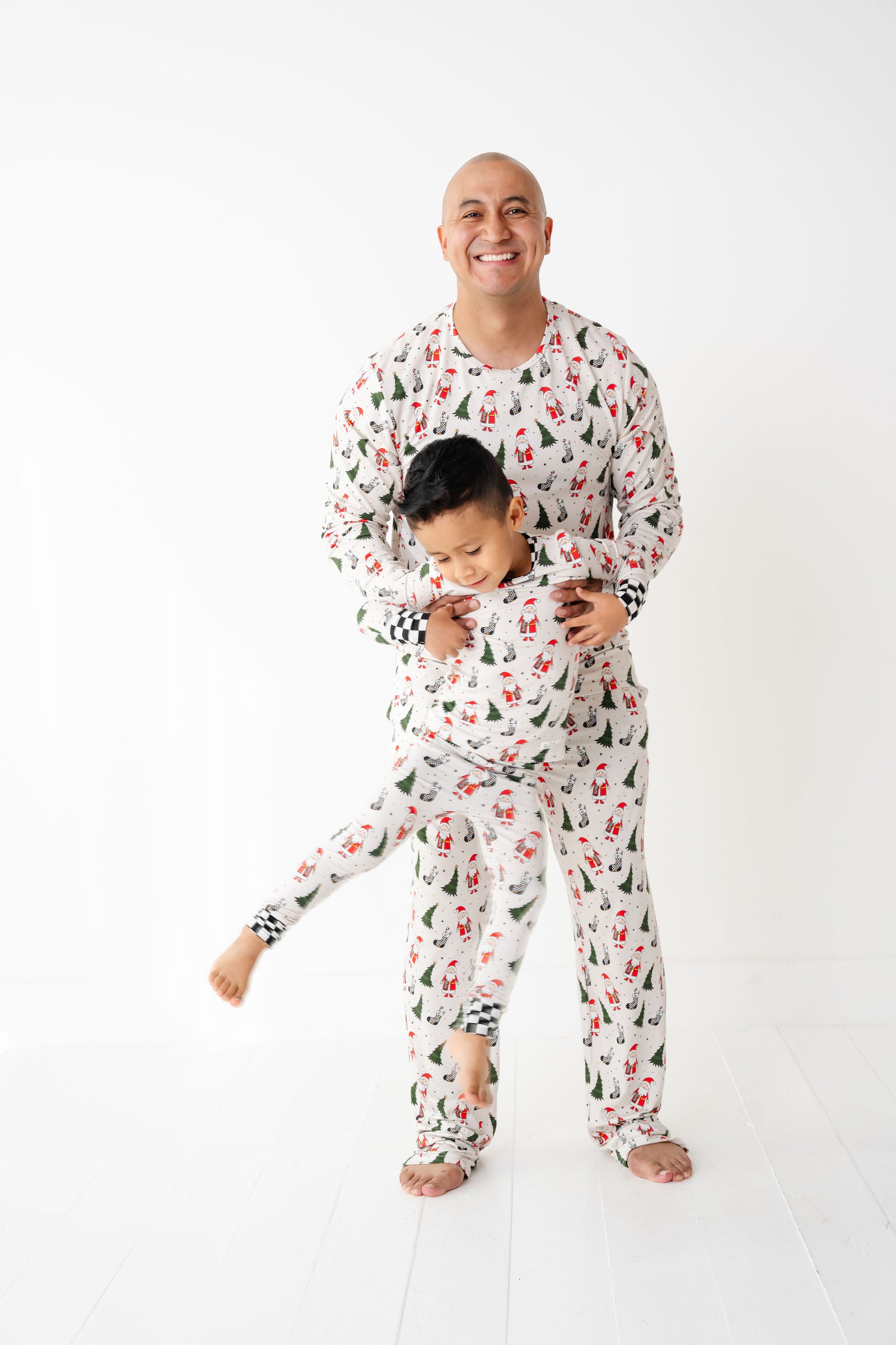 Man and child wearing matching pajamas by Rad toddler called Merry Everything Bamboo Christmas Pajamas with Santa, trees, and checkered stockings