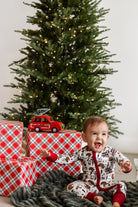A baby wearing a festive white and red Christmas onesie sits on a gray blanket in front of a decorated Christmas tree, with wrapped presents and a small red toy car nearby.