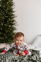 A baby wearing a festive white and red Christmas-themed onesie sits on a cozy green blanket in front of a decorated Christmas tree.