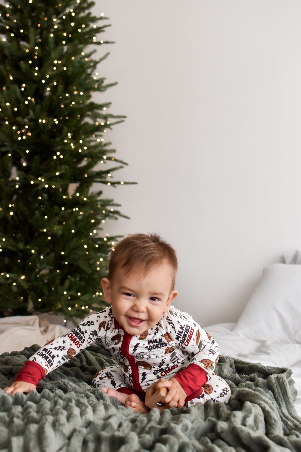 A baby wearing a festive white and red Christmas-themed onesie sits on a cozy green blanket in front of a decorated Christmas tree.