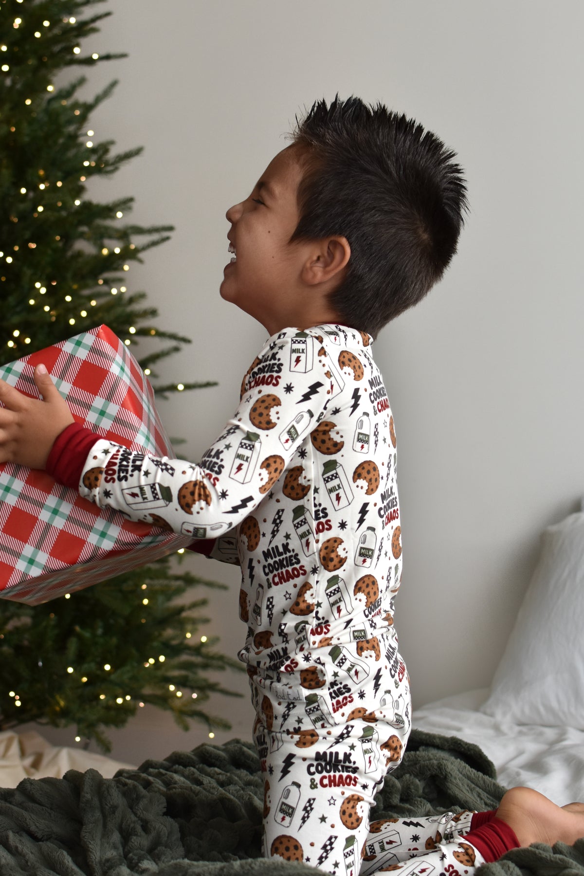 A young boy wearing a white Christmas-themed pajama set with milk cookies and chaos text, sitting on a dark green blanket next to a lit Christmas tree and holding a wrapped gift.