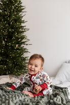 A baby sitting on a bed wearing a festive holiday-themed onesie with a white background, red trim, and a print of gingerbread cookies and reindeer. A decorated Christmas tree and a gray knit blanket are visible in the background.