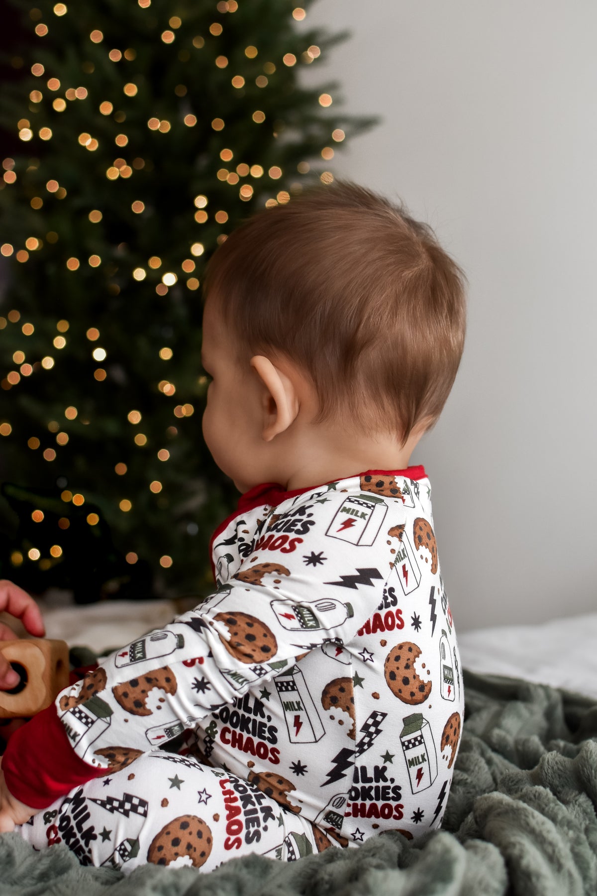 A baby wearing a white Christmas-themed cookie print pajama set, sitting on a gray blanket in front of a glowing Christmas tree.