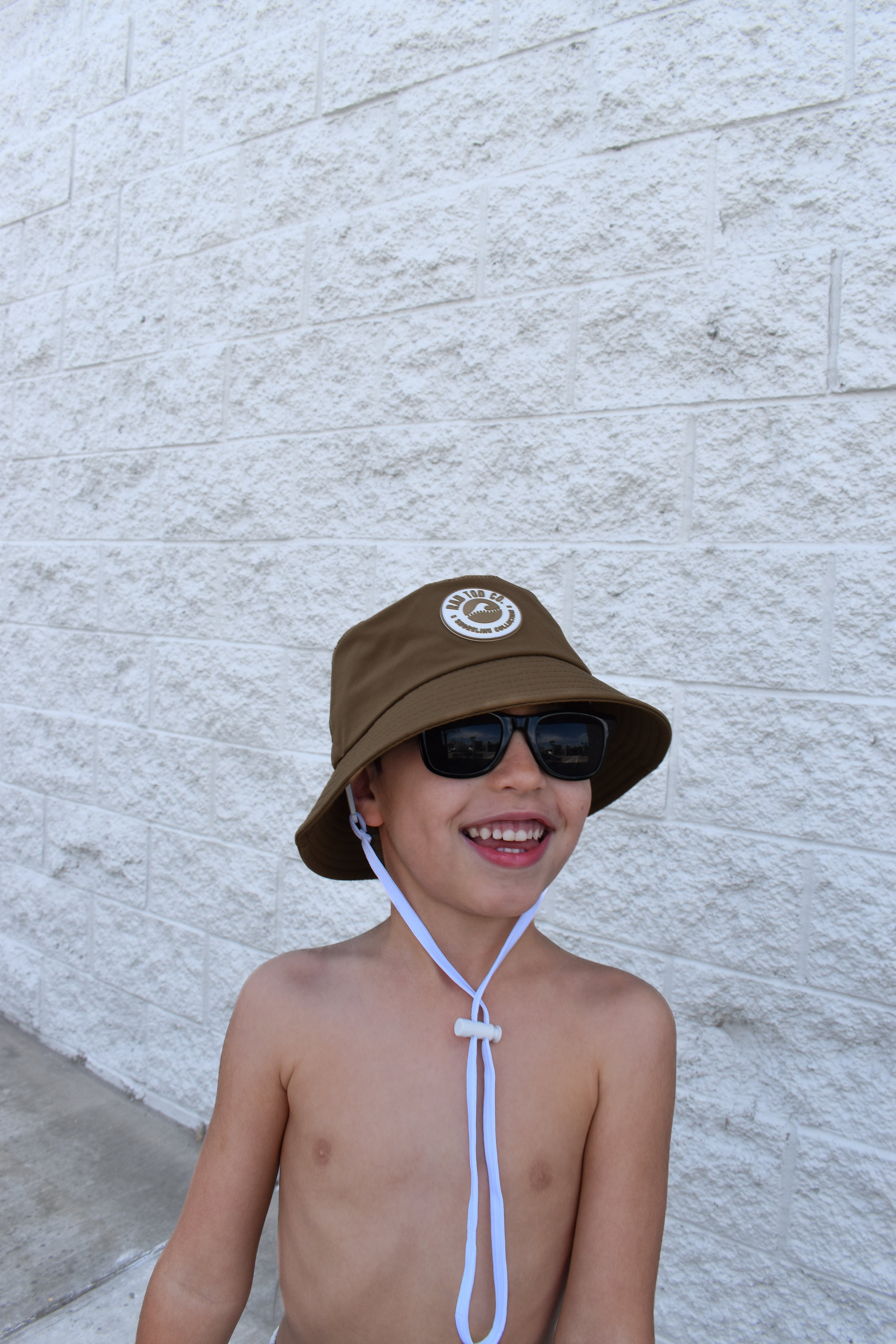 Child wearing a brown bucket hat and sunglasses against a light gray wall.