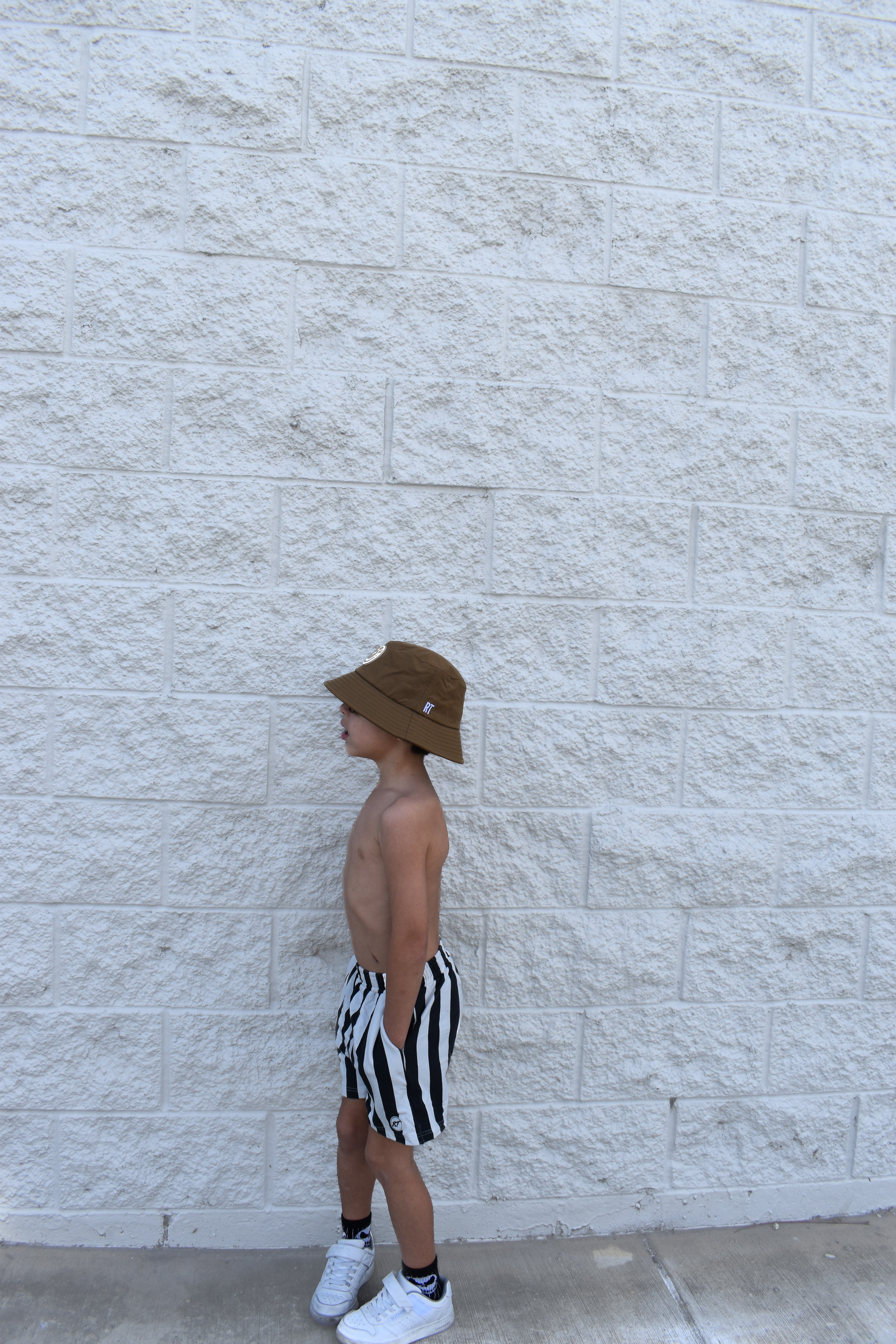 Person wearing a brown bucket hat and black and white striped shorts standing against a light stone wall.