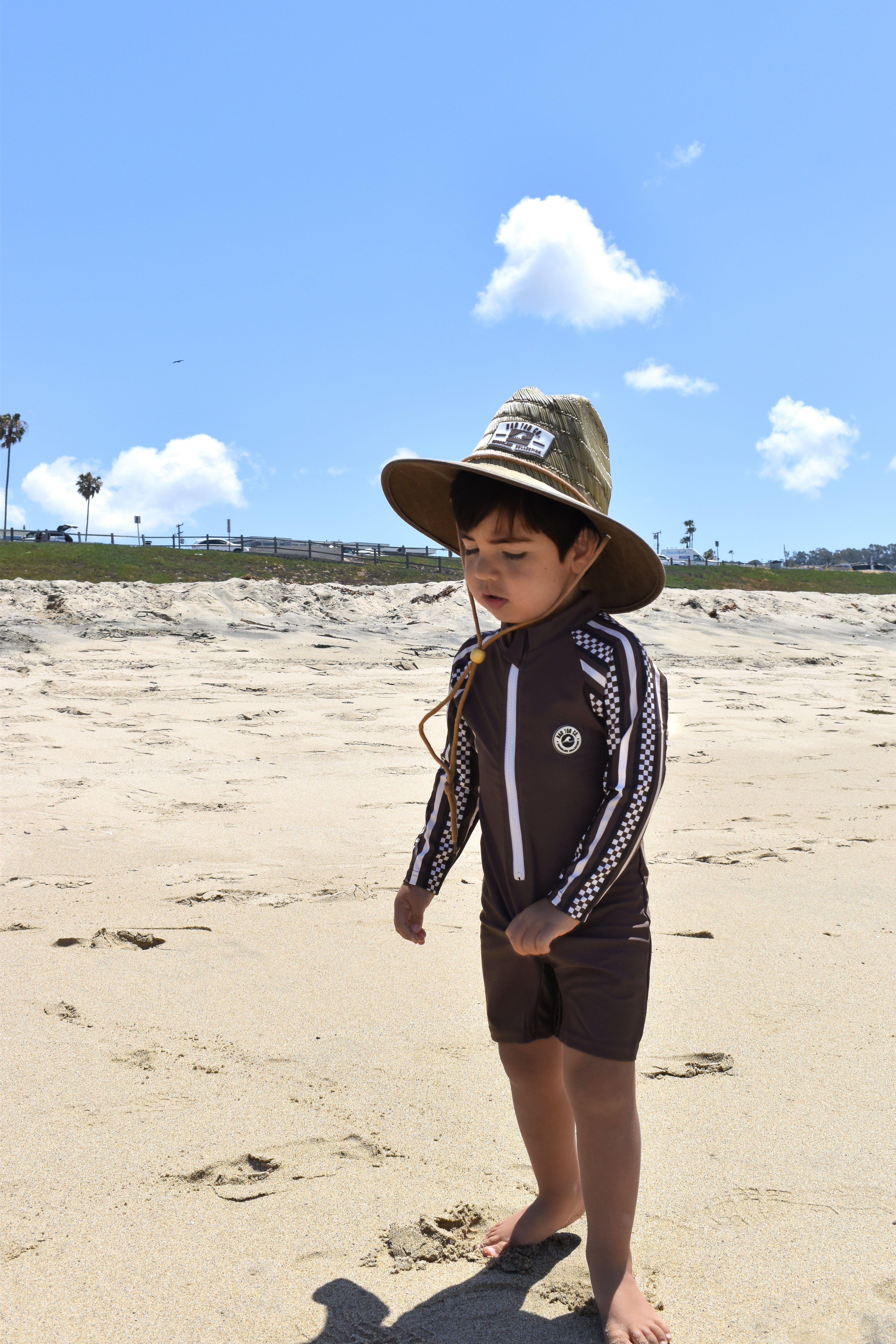 Child wearing a hat and swimsuit on a sandy beach with a clear blue sky.