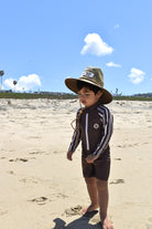 Child wearing a hat and swimsuit on a sandy beach with a clear blue sky.