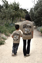 A woman and a young boy walk hand-in-hand on a dirt path, viewed from behind. Both are wearing matching beige graphic t-shirts with black text and coffee cup illustrations.