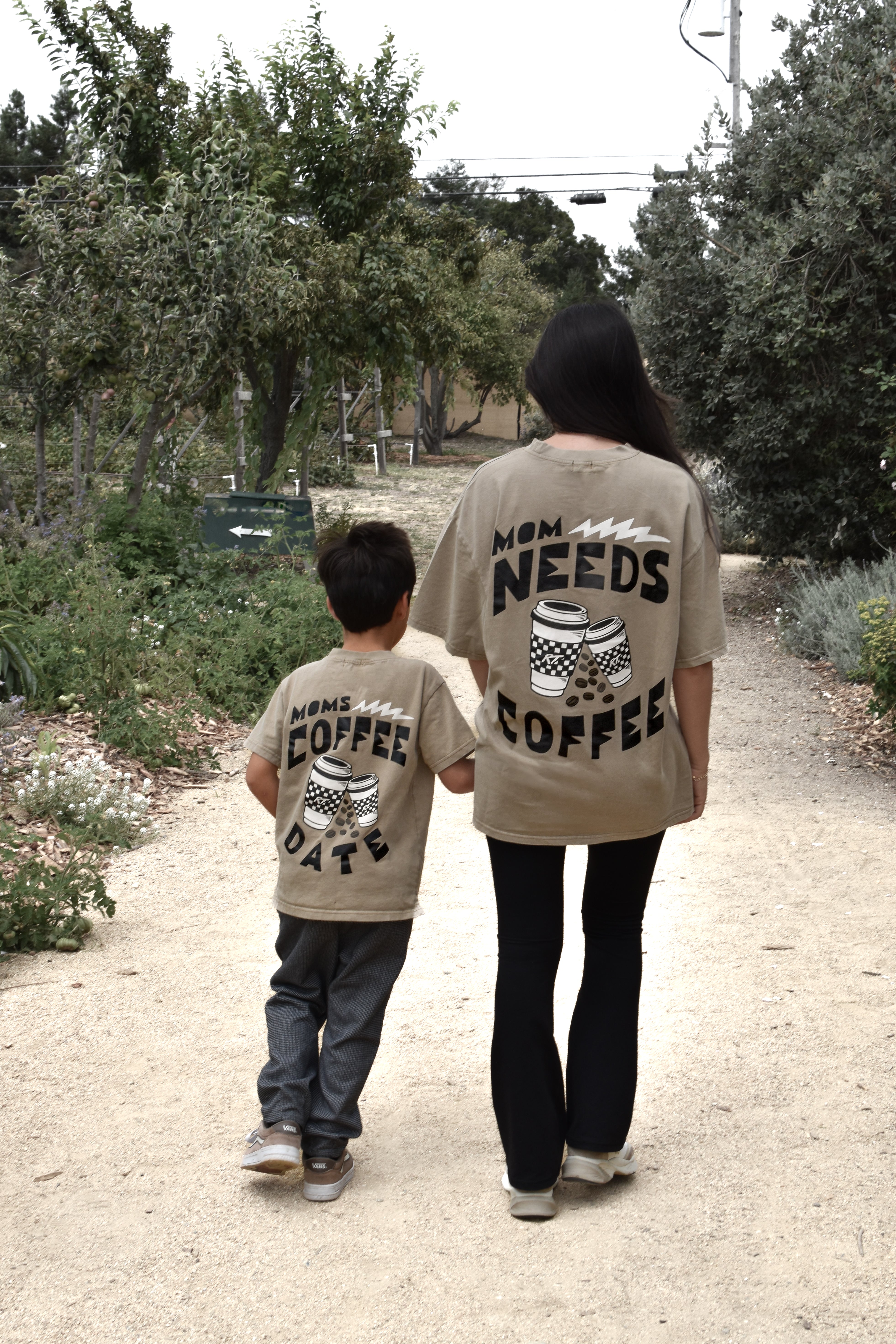 A woman and a young boy walk hand-in-hand on a dirt path, viewed from behind. Both are wearing matching beige graphic t-shirts with black text and coffee cup illustrations.