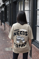 A woman viewed from behind wearing an oversized beige graphic t-shirt with the text 'MOM NEEDS COFFEE' and coffee cup illustrations on the back, standing on a brick sidewalk next to a building with black-framed windows.