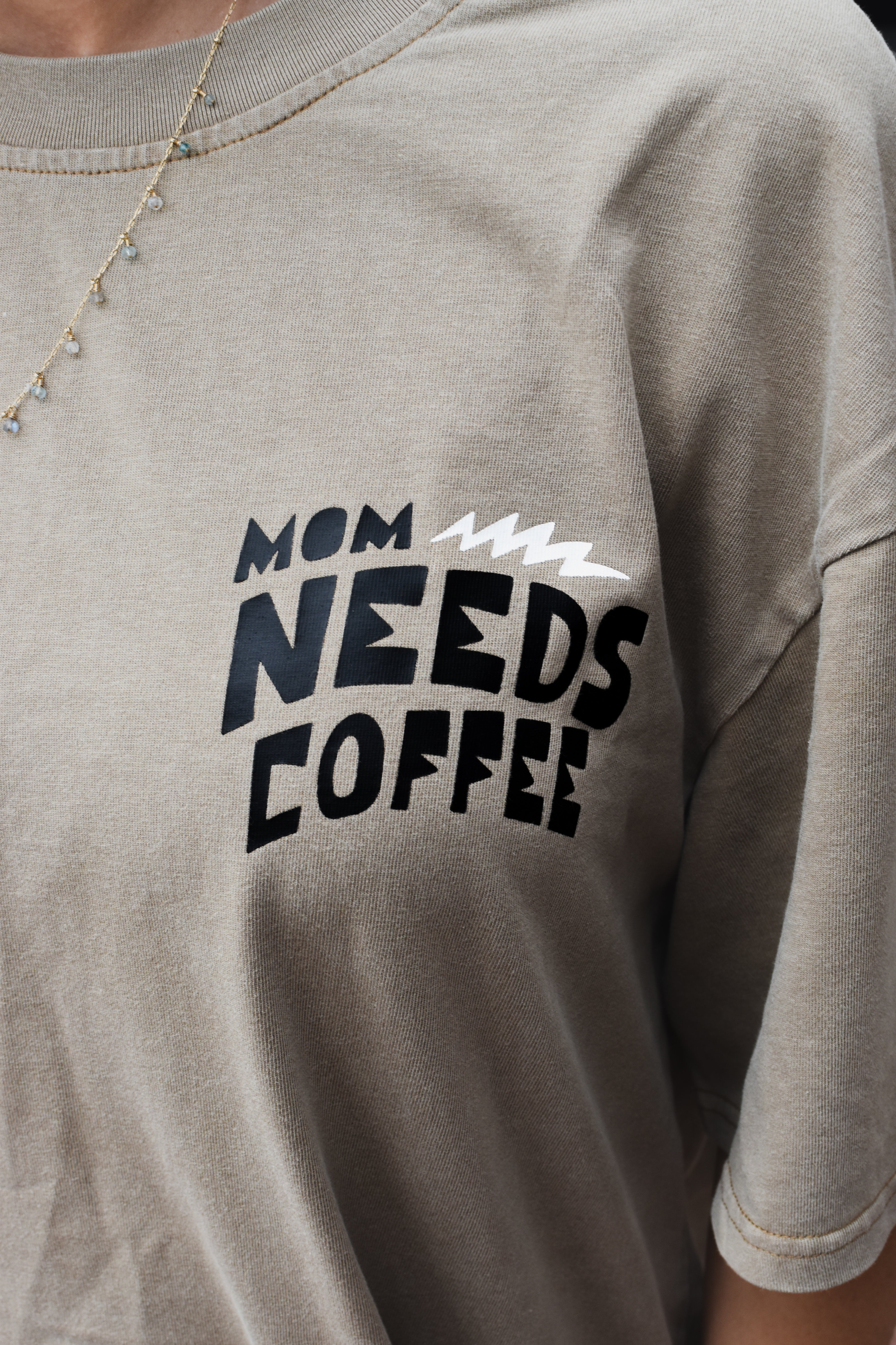 Close-up of a person wearing beige  t-shirt with a black and white graphic that reads 'MOM NEEDS COFFEE'.