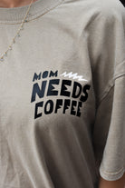 Close-up of a person wearing beige  t-shirt with a black and white graphic that reads 'MOM NEEDS COFFEE'.