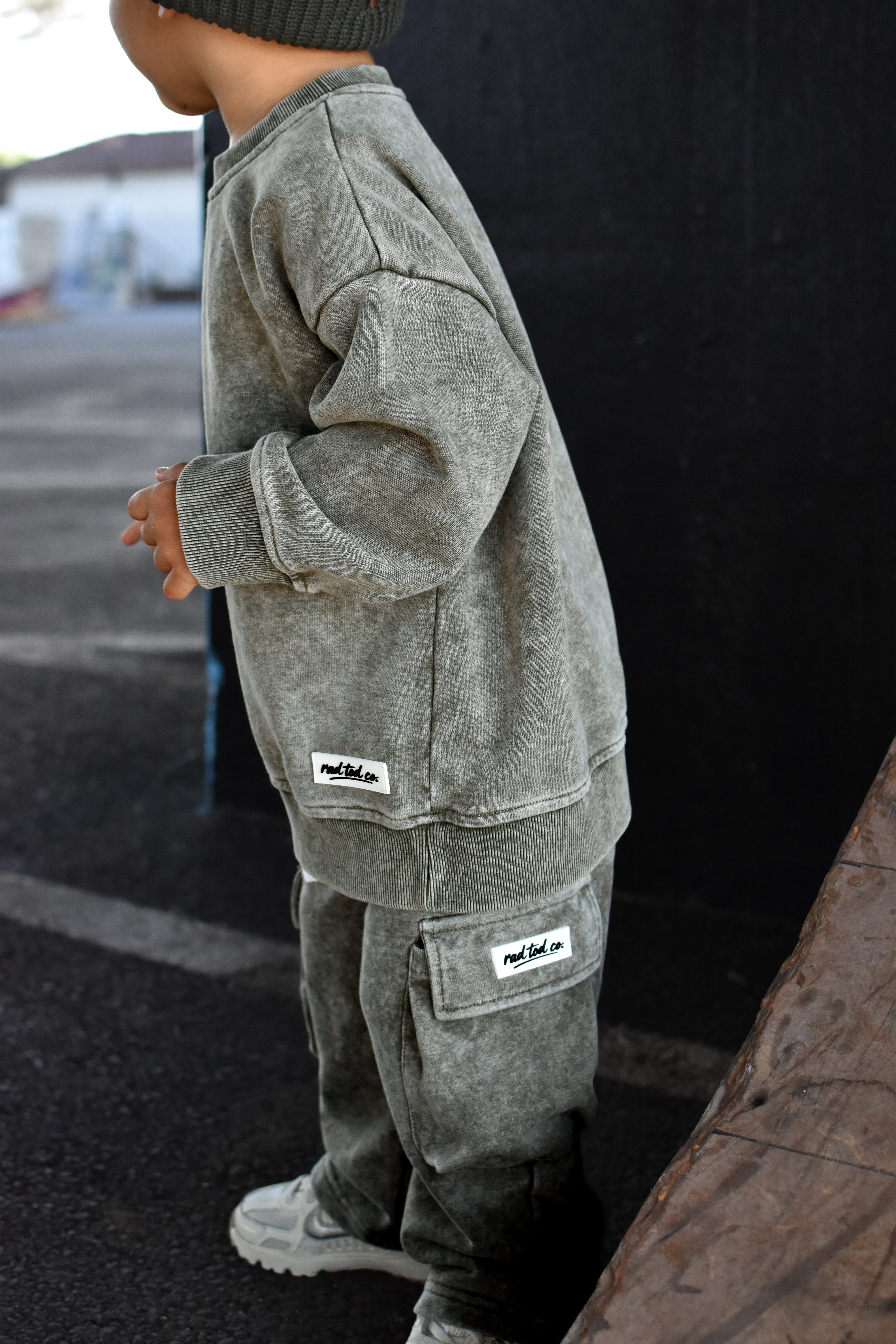 A child wearing a matching moss washed sweatshirt and jogger pants set with small white brand patches, standing outdoors against a dark wall.